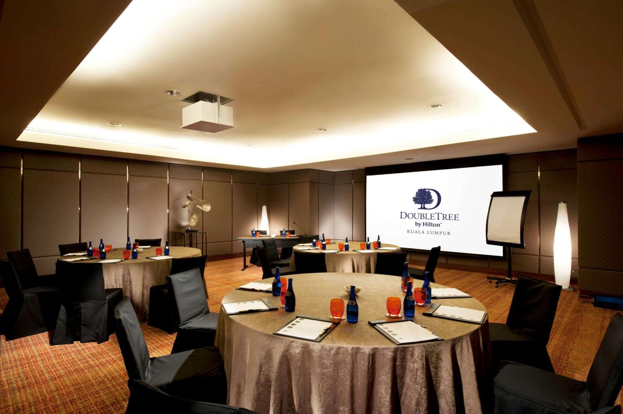 Doubletree By Hilton Kuala Lumpur Ξενοδοχείο Εξωτερικό φωτογραφία The photo shows a conference room set up for a meeting or event. There are several round tables with beige tablecloths, each adorned with blue and red decorations, likely drinks and supplies. Black chairs are placed around the tables. On one wall, th